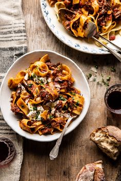 two plates of pasta and bread on a wooden table with utensils next to them