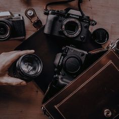 a person holding a camera next to two other cameras on a wooden table with leather cases