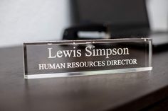 a glass plaque that says lewis simpson human resources director sits on a desk in front of a laptop