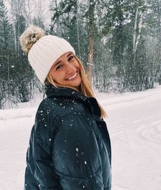 a woman standing in the snow wearing a white hat and black jacket with a pom - pom on it