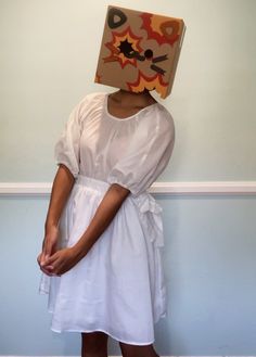 a woman wearing a box on her head standing in front of a wall with a cardboard box on her head