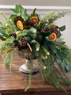 an arrangement of greenery and oranges in a silver vase on a wooden table