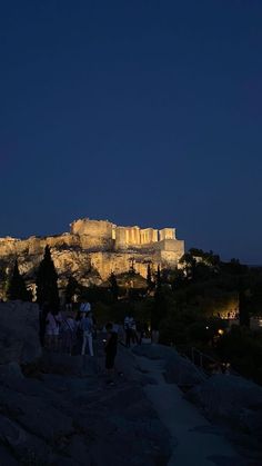 people are standing on the side of a hill at night with an illuminated castle in the background