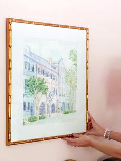 a woman is holding up a painting in front of a pink wall with gold trim