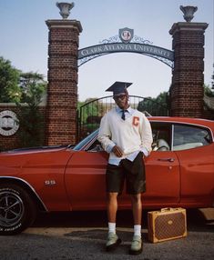 a man standing in front of an orange car wearing a graduation cap and gown with his hands on his hips