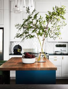 a kitchen with a wooden table topped with a bowl of fruit and a vase filled with green leaves