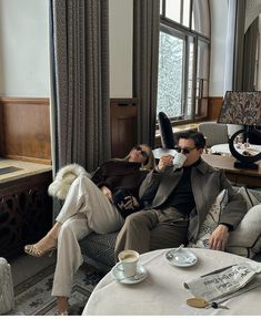 two people sitting on couches drinking coffee in a living room next to a window