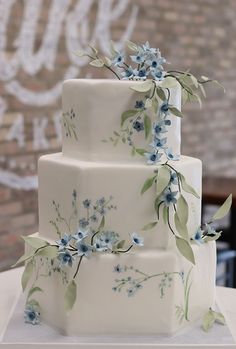 a three tiered cake with blue flowers and leaves on the top is sitting on a table in front of a brick wall