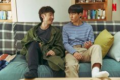 two men sitting on a couch talking to each other in front of bookshelves