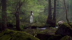 a woman standing in the middle of a forest with moss growing on it's rocks