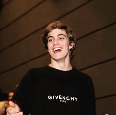 a young man smiling and holding a cell phone in his hand while wearing a givenchy t - shirt