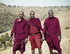 three men in red and purple clothing standing on a dirt road with hills in the background