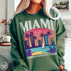 "Celebrate \"Vice City\", the capital of South Florida, the gem of Florida's Golden Coast, the Sunshine City.... MIAMI FLORIDA, with our one of kind Triangle Threads Shop Miami Sweatshirt. This crewneck is perfect for those cold nights on the beach, or warming up after a dip in the pool back at the hotel. Celebrate your bachelorette, your family trip, or a Florida family reunion with this unisex sweatshirt perfect for all styles and ages. The Gildan 18000 50/50 blend fabric has that comfy, lived-in feel, that's like a hug every time you put it on! Now you can take a bit of Miami with you wherever you go! Grab one today! * Product Highlights * Ideal for any situation, this Gildan 18000 heavy blend crewneck sweatshirt is pure comfort. Made from an extremely soft 50/50 polyester and cotton bl Miami Beach Tshirt, Bebesota Sweater, Florida Sweatshirt, Beach Crewneck, Miami Bachelorette, Sunshine City, Miami Vacation, Golden Coast, Beach Sweatshirt