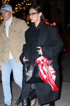 a woman in black coat carrying a red and white bag while standing next to a man