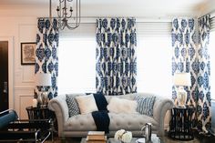 a living room with blue and white curtains on the window sill, two couches, coffee table, lamp, and chandelier