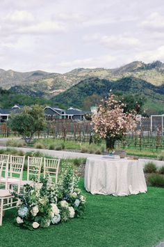 an outdoor ceremony setup with white chairs and flowers on the grass in front of mountains