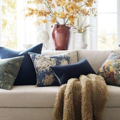 a white couch with many pillows on it in front of a vase and window sill