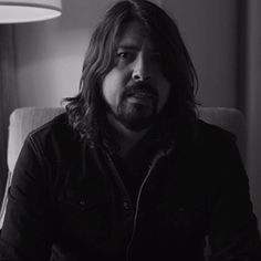 a man with long hair sitting in a chair next to a lamp and looking at the camera