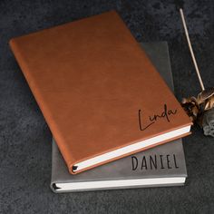 a brown leather book sitting on top of a gray book next to a dead flower