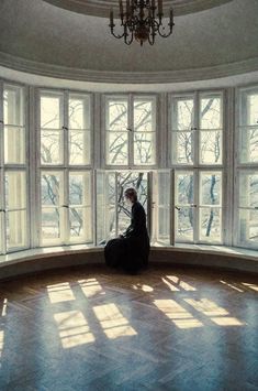 a woman sitting on the floor in front of a large window looking out at trees