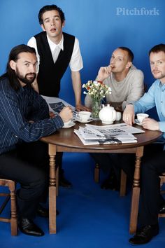 three men sitting at a table with cups and saucers in front of them, posing for the camera