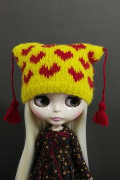 a doll wearing a yellow knitted hat with red hearts and tassels on it