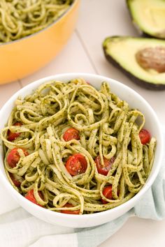 a white bowl filled with pesto pasta next to an avocado on the side