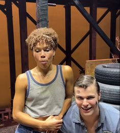 two people standing next to each other in front of stacked tires and one person sticking out her tongue