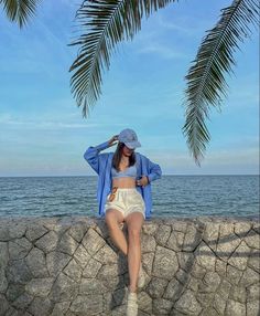 a woman sitting on top of a stone wall next to the ocean wearing a hat