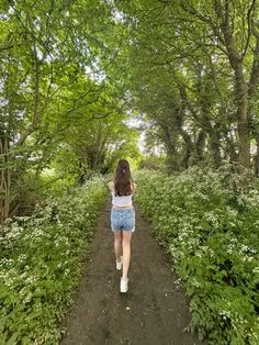 a woman walking down a dirt road surrounded by trees and plants on both sides of the path