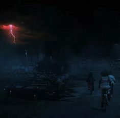 two bicyclists ride down a dark road at night with lightning in the background