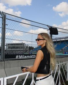 a woman in black top and white pants standing next to fence with stadium behind her