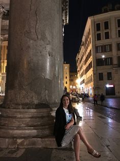 a woman sitting on the side of a building next to a tall pillar at night