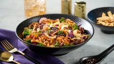 two black bowls filled with pasta and vegetables next to silverware on a gray table