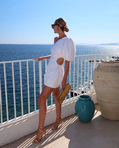 a woman in a white dress standing on a balcony next to the ocean holding a purse