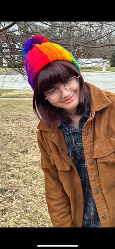 a young woman wearing glasses and a colorful knitted hat stands in front of a tree