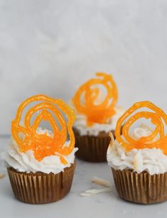 three cupcakes with frosting and orange swirl decorations on top, sitting on a white surface