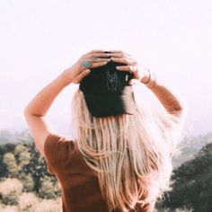 a woman with long blonde hair is holding her hat over her head and looking into the distance