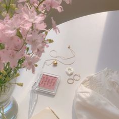 pink flowers in a vase and jewelry on a white table next to an open book