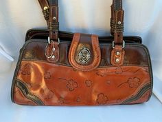 This shoulderbag is a caramel color on the bottom half with a brown color top, sides and bottom of bag. The strap drop is 12.5".  They are brown on one side and caramel on the other side.  There are etched silver hardware holding them on to bag. A zipper closes the purse and there is also a flap with a snap closure. The inside has a satiny lining with 3 compartments. 1 is open with 2 slip pockets 1 is zipped 1 is open with a zipped pocket There are shamrocks imprinted in the outside fa Vintage Brown Bags With Silver-tone Hardware, Retro Brown Satchel With Removable Pouch, Brown Satchel Shoulder Bag With Silver-tone Hardware, Brown Satchel With Silver-tone Hardware For Travel, Cognac Satchel Shoulder Bag With Silver-tone Hardware, Cognac Shoulder Bag With Silver-tone Hardware For Everyday, Brown Leather Satchel With Double Handle, Brown Double Handle Satchel With Leather Backing, Brown Crossbody Bag With Silver-tone Hardware