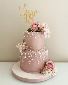 a three tiered cake with pink flowers and gold lettering on top, sitting on a white plate