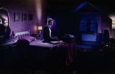 a man sitting on top of a bed in a bedroom next to a dresser and mirror