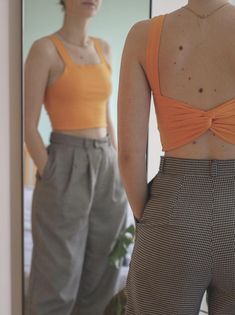 two women standing in front of a mirror with their backs turned to the camera, one wearing an orange tank top