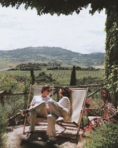 two people sitting on chairs in the middle of an outdoor area with flowers and greenery