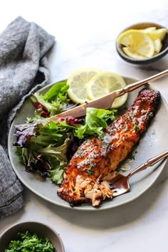 a white plate topped with salmon next to a bowl of greens and lemon wedges