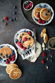 pancakes with berries and blueberries are on plates next to silverware, forks and spoons