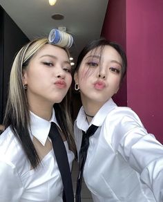 two young women are taking a selfie in front of a pink wall and one is wearing a black tie