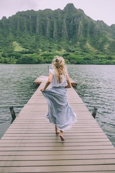 a woman is walking on a dock near the water with mountains in the back ground