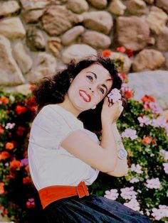 a woman sitting on the ground with flowers in her hand and holding a flower up to her face