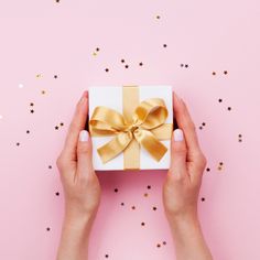 two hands holding a white gift box with gold ribbon on pink background surrounded by confetti